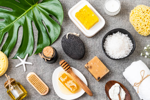 Top view of various skin a body care products such as a bath sponge, a candle, a brush, handmade soaps, honey, a pumice stone, salt, a towel, and a white bottle. Predominant colors are grey, yellow and beige. Studio shot taken with Canon EOS 6D Mark II and Canon EF 24-105 mm f/4L