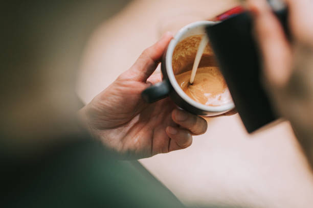 over the shoulder asian chinese male barista pouring froth milk on coffee cup prepared coffee latte art at bar counter