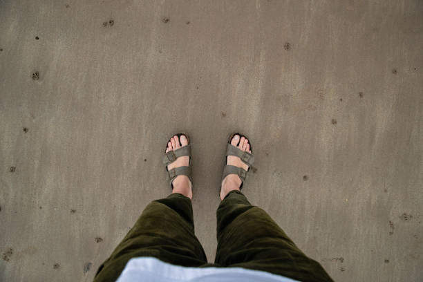 A personal perspective of an unrecognisable person standing on the sand at Beadnell beach, North East England. He is wearing sandals without socks.