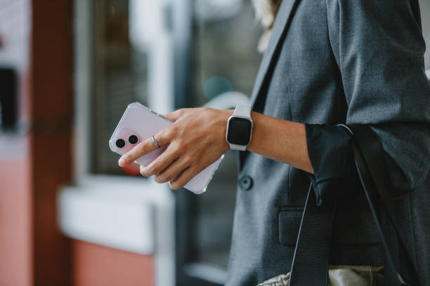 Close-up of a businesswoman holding a smartphone and smartwatch, standing in a city street. Professional attire and modern technology showcase a busy urban lifestyle.