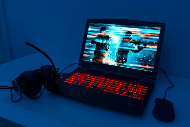 Gaming laptop stands on a table. It is night and the keyboard lights up with red colors. A computer mouse is connected and also headphone with a microphone. The screen shows a video game with a soldier firing a rifle. The image on the screen is the photographer's work